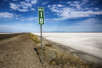 A green mile marker sign reading 'Mile 18' stands near a barren landscape with an expansive salt flat under a bright blue sky. The road runs parallel to the flat, bordered by dry shrubs and earth.