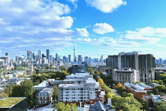 A vibrant cityscape featuring a skyline filled with modern buildings and a prominent tower in the distance. The scene captures a variety of architectural styles, from residential houses to high-rise skyscrapers. The sky is mostly clear with some fluffy white clouds, and greenery is interspersed between the buildings.
