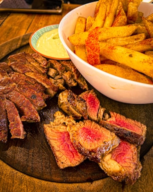 A wooden platter displays slices of juicy, medium-rare steak alongside a bowl of seasoned French fries. A small dish of creamy dipping sauce accompanies the dish, enhancing the savory presentation.