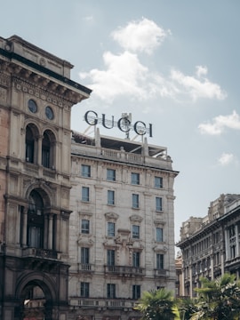 A grand and ornate historical building with classic architectural features like arched windows and decorative stonework. At the top of the building, a large sign with the text 'GUCCI' is prominently displayed. The sky above is clear and blue with several fluffy white clouds. Some palm trees are visible in the foreground, adding a touch of greenery.