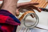 A person applies a transparent label with decorative deer graphics onto a glass jar. The scene shows hands carefully placing the label on the jar, with a red and blue plaid shirt visible. Various items, including black and red pens, are scattered on the table.