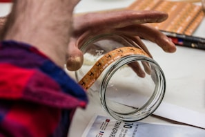 A person applies a transparent label with decorative deer graphics onto a glass jar. The scene shows hands carefully placing the label on the jar, with a red and blue plaid shirt visible. Various items, including black and red pens, are scattered on the table.