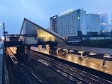 A dimly lit train station platform with tracks running underneath an overpass, featuring the name 'Warszawa Ochota' on the side. The platform is illuminated by overhead lights, and a few people are visible walking or waiting. In the background, there is a large hotel building with the name 'Campanile' and other advertisements. The overall atmosphere is calm with a slightly overcast sky.