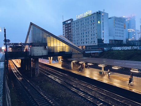 A dimly lit train station platform with tracks running underneath an overpass, featuring the name 'Warszawa Ochota' on the side. The platform is illuminated by overhead lights, and a few people are visible walking or waiting. In the background, there is a large hotel building with the name 'Campanile' and other advertisements. The overall atmosphere is calm with a slightly overcast sky.
