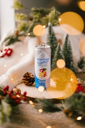 A beverage can labeled 'Positive Beverage' sits on a wooden surface surrounded by festive decorations, including pinecones, evergreen branches, and red berries. Soft, warm lighting with circular bokeh effects adds a cozy, holiday ambiance.