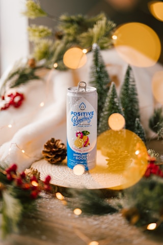 A beverage can labeled 'Positive Beverage' sits on a wooden surface surrounded by festive decorations, including pinecones, evergreen branches, and red berries. Soft, warm lighting with circular bokeh effects adds a cozy, holiday ambiance.