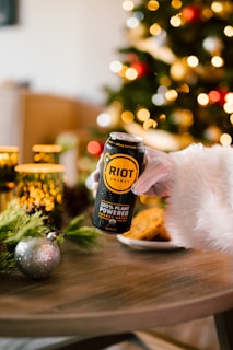 A hand wearing a white fur sleeve holds a can of Riot Energy drink over a wooden table adorned with festive decorations, such as pine branches and a shiny silver ornament. In the background, there is a blurred view of Christmas tree lights and part of a tree.