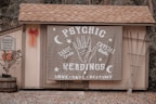 A small wooden structure with a shingled roof displays a large banner advertising psychic readings with elements like a hand, moons, and stars. To the left, a sign denotes reserved parking for a 'mystical journey' alongside a potted plant with orange flowers and a red bow on the wall.