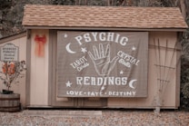 A small wooden structure with a shingled roof displays a large banner advertising psychic readings with elements like a hand, moons, and stars. To the left, a sign denotes reserved parking for a 'mystical journey' alongside a potted plant with orange flowers and a red bow on the wall.
