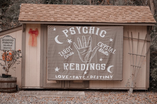 A small wooden structure with a shingled roof displays a large banner advertising psychic readings with elements like a hand, moons, and stars. To the left, a sign denotes reserved parking for a 'mystical journey' alongside a potted plant with orange flowers and a red bow on the wall.
