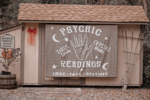 A small wooden structure with a shingled roof displays a large banner advertising psychic readings with elements like a hand, moons, and stars. To the left, a sign denotes reserved parking for a 'mystical journey' alongside a potted plant with orange flowers and a red bow on the wall.