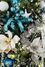 Various Christmas ornaments adorn a tree, including a white flower with glittery gold accents, blue baubles, and silver snowflakes. The decorations feature shimmering materials and intricate designs, set against a backdrop of lush green pine needles.