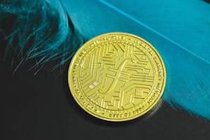 A gold coin with intricate circuit-like patterns rests on a surface with vivid blue feathers to its side. The coin carries inscriptions and a distinct currency symbol, emphasizing a digital or futuristic theme.