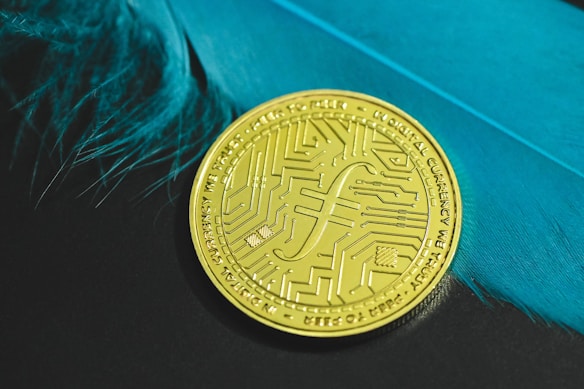 A gold coin with intricate circuit-like patterns rests on a surface with vivid blue feathers to its side. The coin carries inscriptions and a distinct currency symbol, emphasizing a digital or futuristic theme.