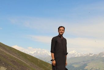 A man stands on a hilltop, dressed in traditional attire. The background features a breathtaking view of green hills and snow-capped mountains under a clear blue sky.