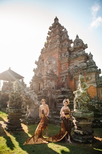 A traditional Balinese temple with intricate stone carvings is set against a bright sky. Two individuals dressed in ornate Balinese costume stand in front of the temple, posing elegantly. The lush greenery around them adds to the vibrant yet serene atmosphere.