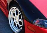 A close-up view of a car featuring a shiny, metallic alloy wheel and a partial view of the vehicle's headlight. The car has a red and black paint job with the black section on the hood area. The smooth, reflective surface of the wheel highlights intricate design and craftsmanship.