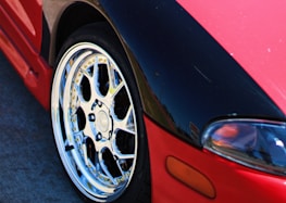 A close-up view of a car featuring a shiny, metallic alloy wheel and a partial view of the vehicle's headlight. The car has a red and black paint job with the black section on the hood area. The smooth, reflective surface of the wheel highlights intricate design and craftsmanship.