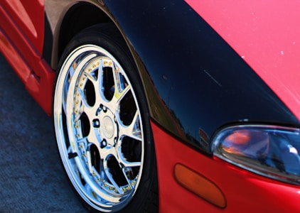 A close-up view of a car featuring a shiny, metallic alloy wheel and a partial view of the vehicle's headlight. The car has a red and black paint job with the black section on the hood area. The smooth, reflective surface of the wheel highlights intricate design and craftsmanship.