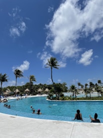A serene and inviting swimming pool surrounded by palm trees and lush greenery, set under a clear blue sky with a few fluffy clouds. People are leisurely enjoying the water, some swimming and others relaxing by the poolside. Straw-roofed huts can be seen in the background, enhancing the tropical ambiance.