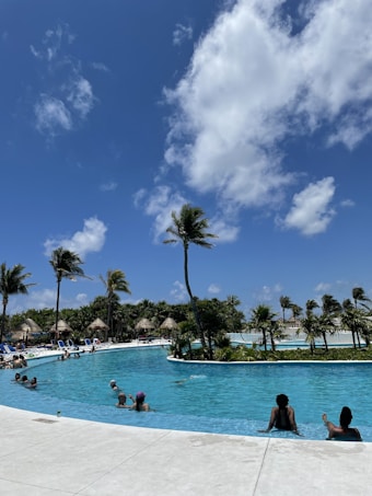 A serene and inviting swimming pool surrounded by palm trees and lush greenery, set under a clear blue sky with a few fluffy clouds. People are leisurely enjoying the water, some swimming and others relaxing by the poolside. Straw-roofed huts can be seen in the background, enhancing the tropical ambiance.