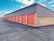 A row of storage units with bright orange roll-up doors and a brick facade. The structure is aligned in a straight row, adjacent to an asphalt driveway. The sky is clear with some clouds, adding a sense of openness to the scene.