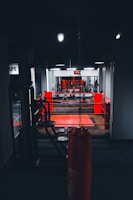 A dimly lit gym with a focus on a boxing ring surrounded by red ropes. In the foreground, there is a hanging red punching bag positioned centrally. The gym equipment is visible along the sides, and a wall clock is seen in the background above the ring, displaying the time. The setting conveys a sense of readiness for physical activity.