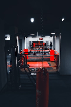 A dimly lit gym with a focus on a boxing ring surrounded by red ropes. In the foreground, there is a hanging red punching bag positioned centrally. The gym equipment is visible along the sides, and a wall clock is seen in the background above the ring, displaying the time. The setting conveys a sense of readiness for physical activity.