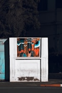 A stylized painting on a white wall features abstract faces with exaggerated features and vibrant colors. The artwork is set against a backdrop of a partially visible building and a shadowy tree. The faces have large blue and black eyes, with prominent lips in shades of red and orange.