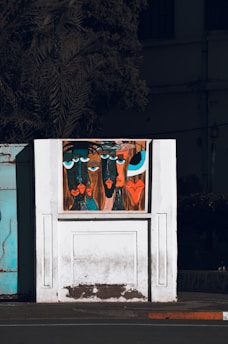A stylized painting on a white wall features abstract faces with exaggerated features and vibrant colors. The artwork is set against a backdrop of a partially visible building and a shadowy tree. The faces have large blue and black eyes, with prominent lips in shades of red and orange.