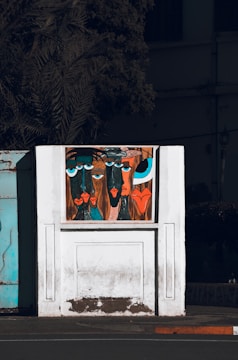 A stylized painting on a white wall features abstract faces with exaggerated features and vibrant colors. The artwork is set against a backdrop of a partially visible building and a shadowy tree. The faces have large blue and black eyes, with prominent lips in shades of red and orange.