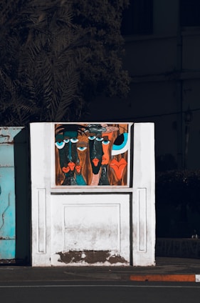 A stylized painting on a white wall features abstract faces with exaggerated features and vibrant colors. The artwork is set against a backdrop of a partially visible building and a shadowy tree. The faces have large blue and black eyes, with prominent lips in shades of red and orange.