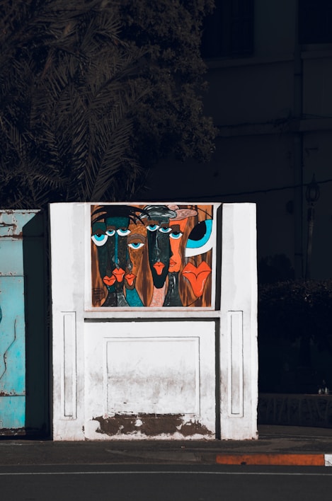 A stylized painting on a white wall features abstract faces with exaggerated features and vibrant colors. The artwork is set against a backdrop of a partially visible building and a shadowy tree. The faces have large blue and black eyes, with prominent lips in shades of red and orange.
