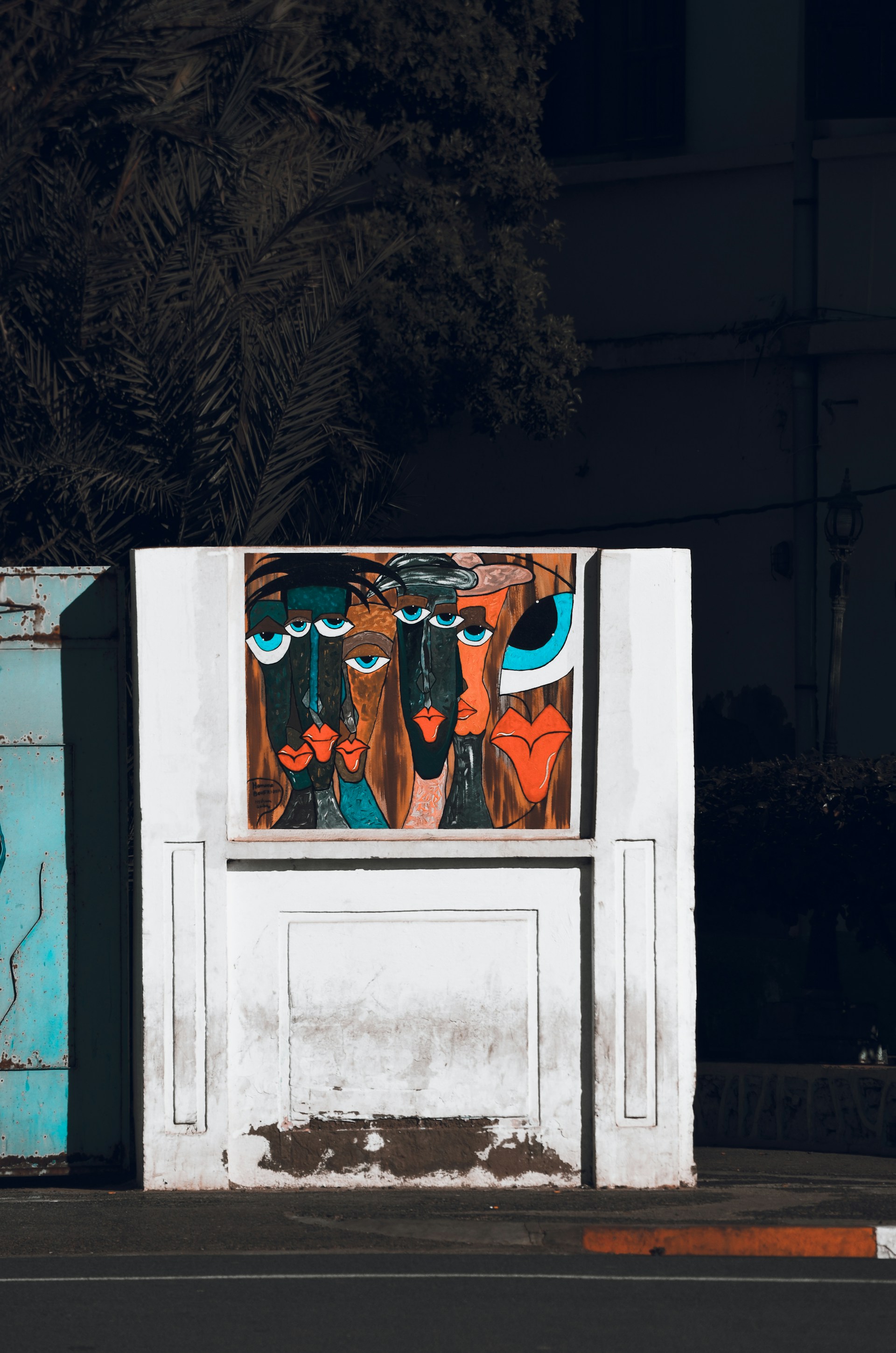 A stylized painting on a white wall features abstract faces with exaggerated features and vibrant colors. The artwork is set against a backdrop of a partially visible building and a shadowy tree. The faces have large blue and black eyes, with prominent lips in shades of red and orange.