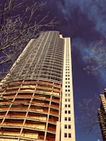 A tall building under construction with a modern architectural style is shown from a low angle. The structure features exposed floors and scaffolding, with metal and glass elements. Leafless tree branches are visible in the foreground, contrasting the deep blue sky.