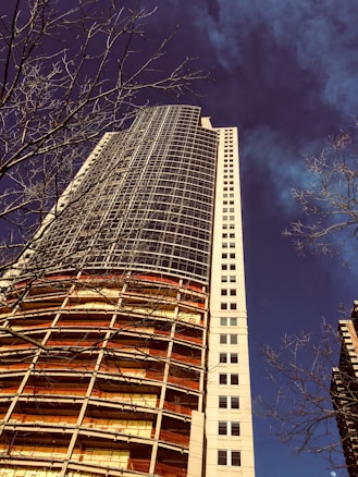 A tall building under construction with a modern architectural style is shown from a low angle. The structure features exposed floors and scaffolding, with metal and glass elements. Leafless tree branches are visible in the foreground, contrasting the deep blue sky.