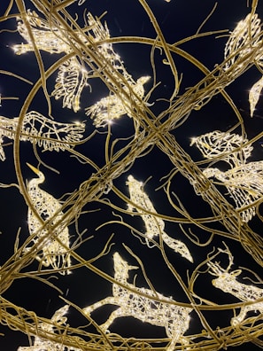 Intricate sculptures made of twinkling lights and metal wires are shaped like various animals, including birds and deer. The structures are intertwined in an elaborate circular pattern against a dark background, creating a mesmerizing and festive appearance.