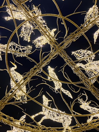 Intricate sculptures made of twinkling lights and metal wires are shaped like various animals, including birds and deer. The structures are intertwined in an elaborate circular pattern against a dark background, creating a mesmerizing and festive appearance.