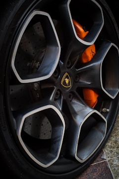 A close-up of a car wheel featuring a stylish rim with a geometric design and a logo at the center. The brake caliper behind the rim is visible in a bright orange color.