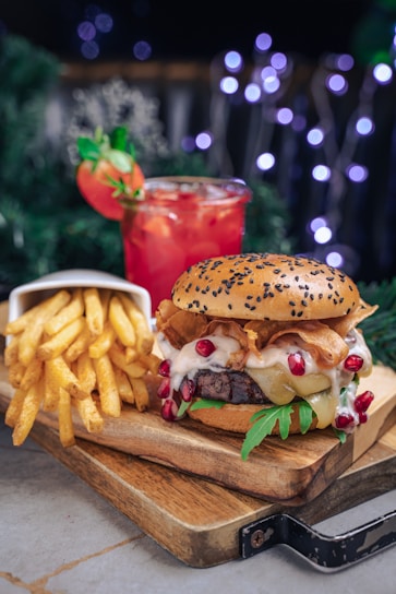 A juicy burger with a sesame seed bun, cheese, bacon strips, lettuce, and pomegranate seeds sits on a wooden board next to a serving of golden French fries in a white container. Behind the burger and fries is a tall glass of a red drink with ice and garnished with a strawberry and a sprig of mint. The background is decorated with bokeh lights creating a festive atmosphere.