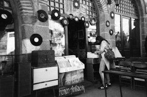 A black and white photo of a person browsing through vinyl records outside a vintage store. Vinyl records are displayed on the stone wall facade of a shop with arched windows. The scene gives a nostalgic feel with visible vintage furniture and decorations.