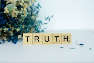 Wooden Scrabble tiles spell out the word 'TRUTH', placed on a surface with a background of dried flowers, including blue and beige tones.