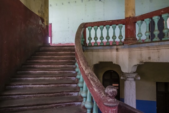 An old, worn staircase with a curving banister is set against a faded, rustic interior. The balustrade features alternating rounded and square shapes, and the walls exhibit peeling paint. Light filters through an open doorway or window, casting soft illumination.