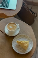 A cup of cappuccino with latte art sits on a saucer on a circular wooden table. Nearby, there is a patterned plate with a slice of yellow cake. A laptop is partially visible on the table, and a brown leather bag is placed on the floor.