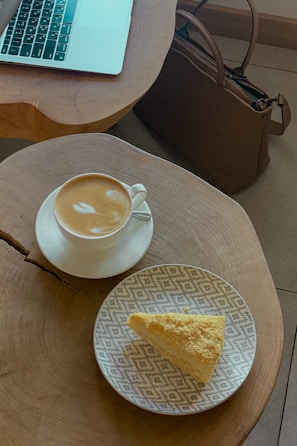 A cup of cappuccino with latte art sits on a saucer on a circular wooden table. Nearby, there is a patterned plate with a slice of yellow cake. A laptop is partially visible on the table, and a brown leather bag is placed on the floor.