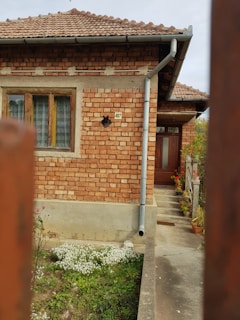 A brick house with a tiled roof and a number 497 address plate next to a window on the side wall. The house features a drainage pipe running along the side wall down to the ground. There is a flower garden at the base of the house with various blooming flowers and plants. The wooden front door is located up a short flight of steps, with potted plants arranged along the railing.