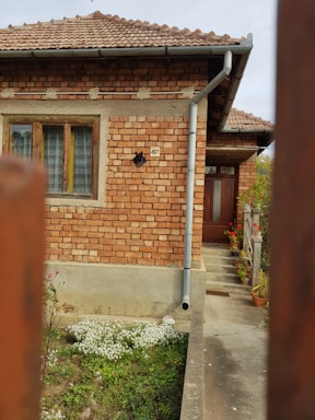 A brick house with a tiled roof and a number 497 address plate next to a window on the side wall. The house features a drainage pipe running along the side wall down to the ground. There is a flower garden at the base of the house with various blooming flowers and plants. The wooden front door is located up a short flight of steps, with potted plants arranged along the railing.