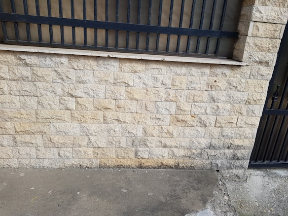 A textured stone wall with a wrought iron fence on top. The wall consists of square stone tiles with variations in color, predominantly beige and light brown. At the bottom, there is a rough concrete surface with visible wear and discoloring. The fence above is composed of vertical black bars, providing a stark contrast to the lighter wall.