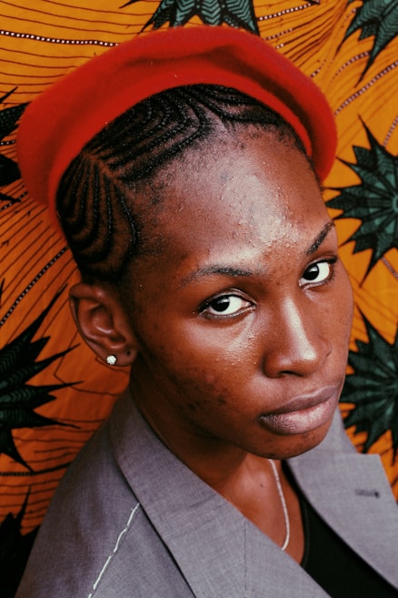 A person with intricately braided hair wearing a red beret and a gray blazer is positioned in front of a vibrant orange fabric with dark green and black patterns. The person's expression is serious, and the lighting highlights their facial features and the texture of the fabric.