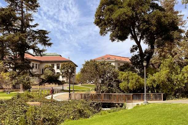 A serene landscape featuring an outdoor academic setting with large trees, lush green grass, and classic architectural buildings with red-tiled roofs. A wooden bridge and a lamp post are visible in the foreground, while people walk along the path, enjoying the sunny day.